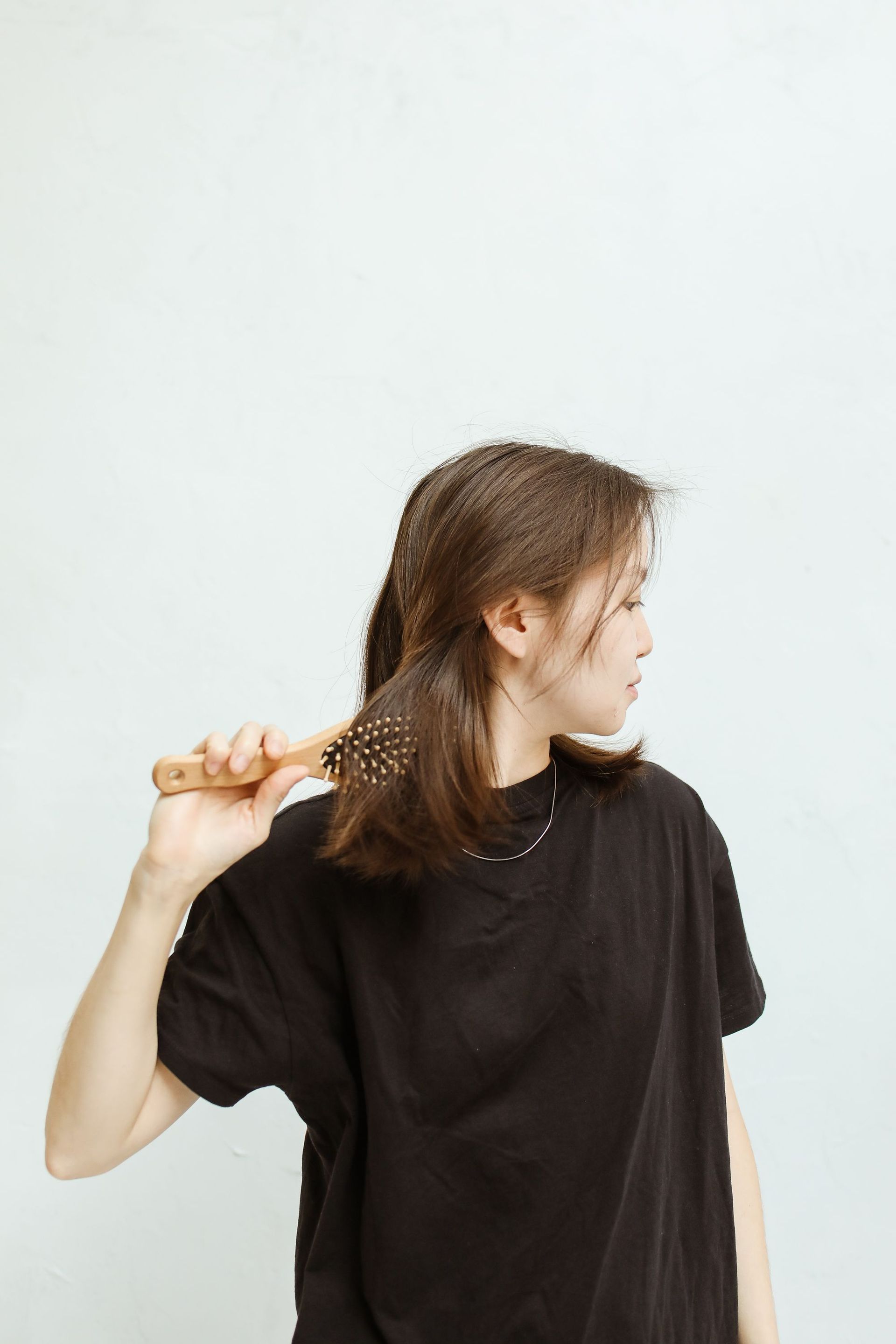 Asian woman combing hair with wooden brush, gazing sideways in simple, minimalist setting.